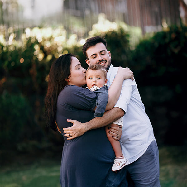 A family of three smiles and hugs each other joyfully in a sunny park setting.